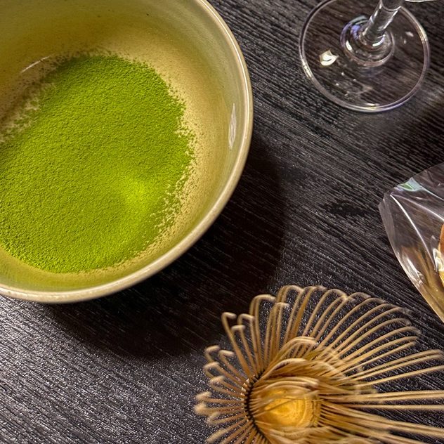 Bol avec du thé matcha, verre à vin et un autre verre sur une table sombre.